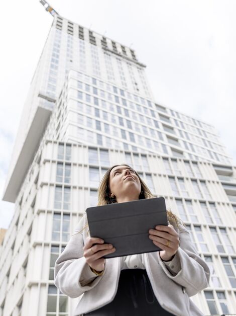 Donna bionda con iPad in mano che guarda verso l’alto in direzione di un edificio residenziale bianco. 